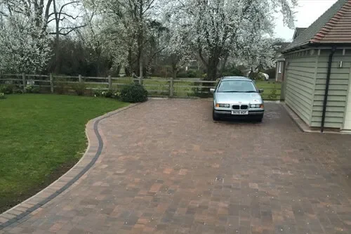 A silver car parked on a brick driveway next to a house, with a wooden fence and trees in the background.