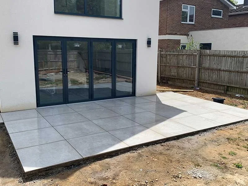 Newly installed gray-tiled patio outside a modern white house with large sliding glass doors and a wooden fence in the backyard.