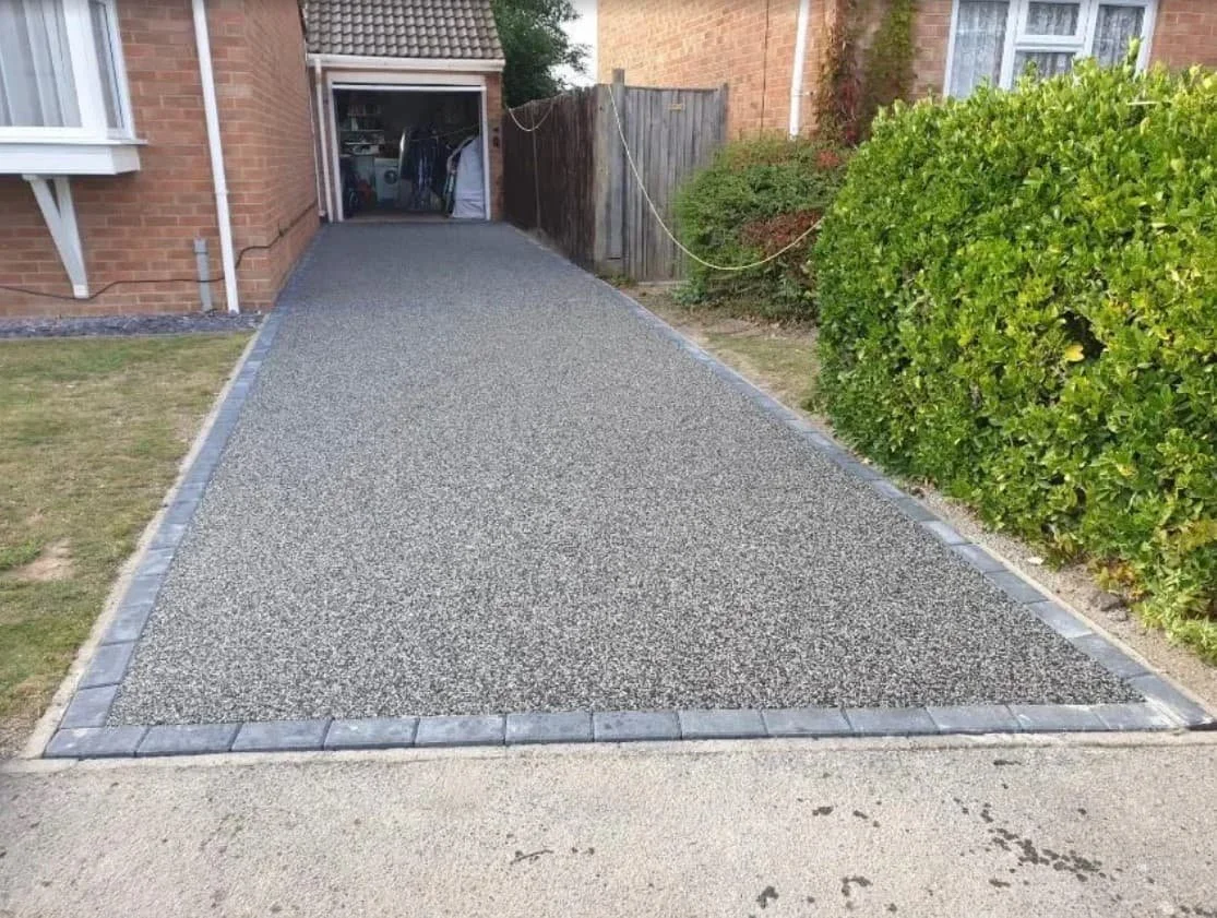 Driveway leading to a garage, bordered by gray bricks, with a house on the left, a wooden fence ahead, and a green hedge on the right.