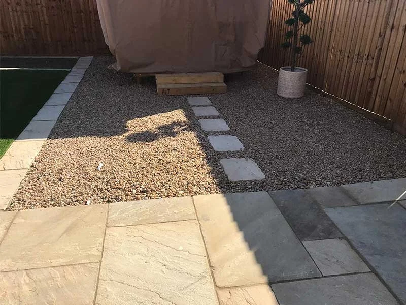 Backyard with a gravel area, stepping stone pathway, covered object on a wooden pallet, potted plant, and a fence.