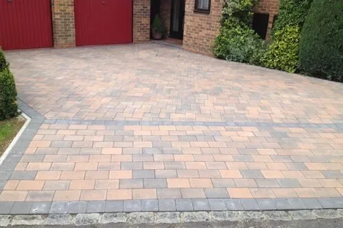 A paved driveway with multicolored bricks in front of a house with a red garage door and surrounded by green shrubs and plants.