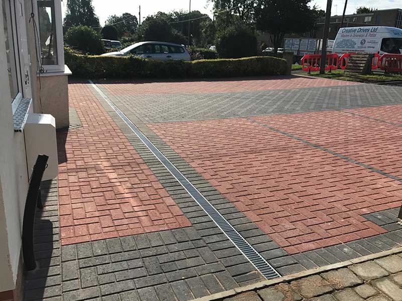 Paved area with red and gray bricks, drainage grate in the center, and cars visible in the background.