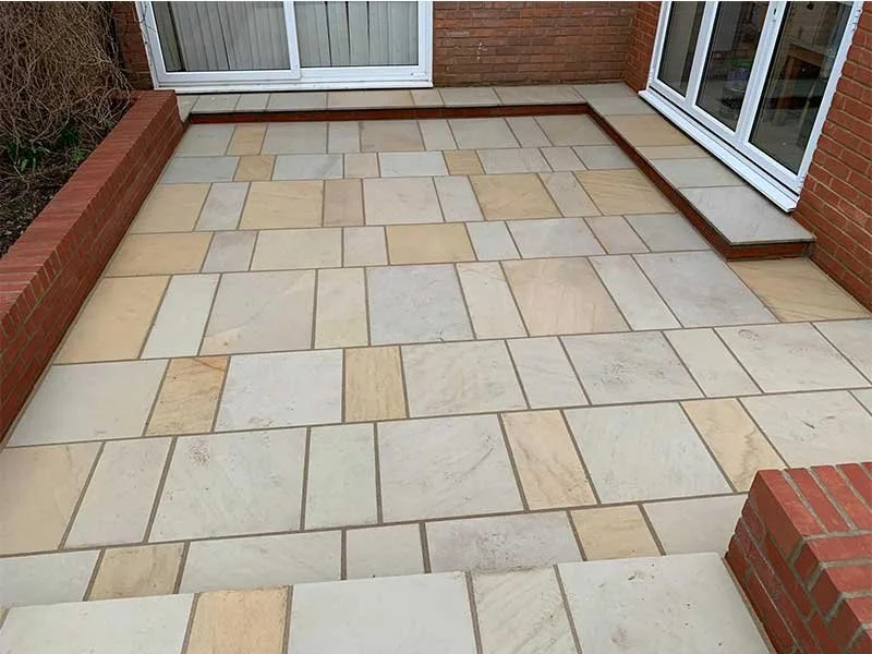 View of a new tiled patio with beige and cream-colored stones, bordered by red brick walls and a house wall with glass sliding doors.