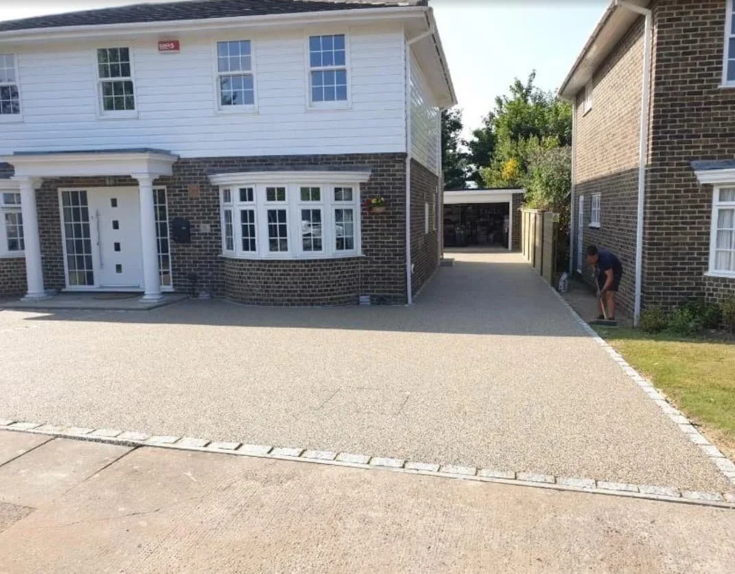 A front yard with a driveway leading to a garage in the background. The house on the left has a brick and white siding facade with large windows and a white front door. The house on the right is brick with several windows. A person is working near th