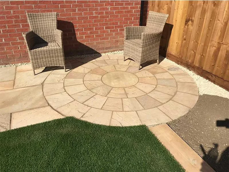 Two wicker chairs with black cushions on a circular stone patio, surrounded by a brick wall, wooden fence, and patches of artificial grass and gravel.