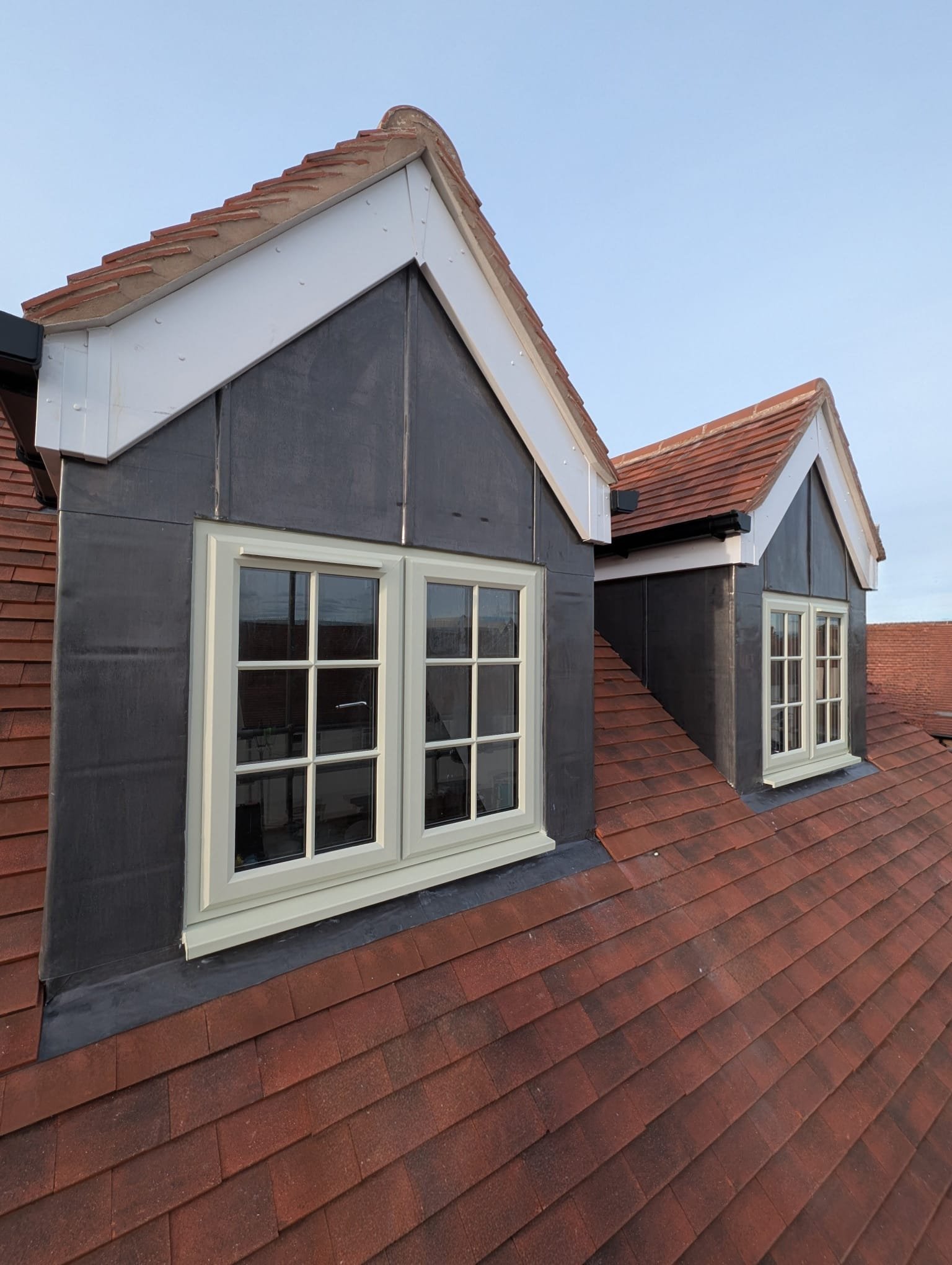 A residential house roof with two dormer windows, red shingles, black flashing, and a blue sky background.