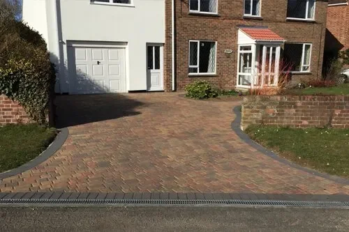 Paved driveway leading to a white garage door and a brick house with a porch, bordered by grass and brick walls.