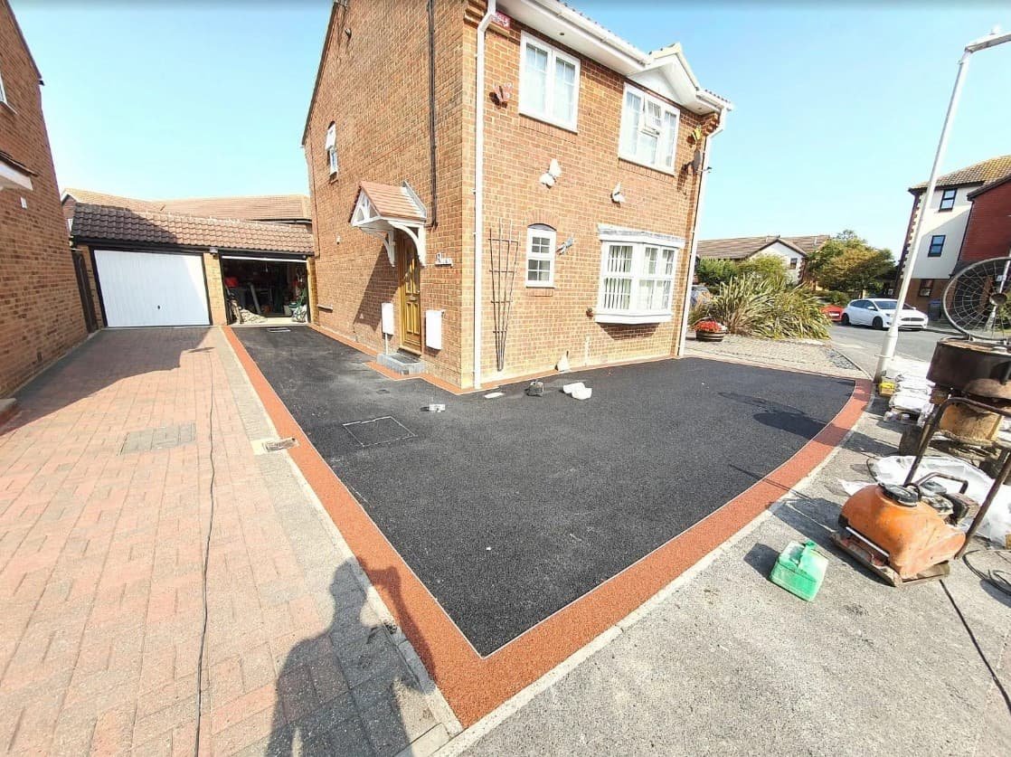 Newly paved black asphalt driveway with a brick border in front of a two-story brick house with white window frames and a wooden front door.