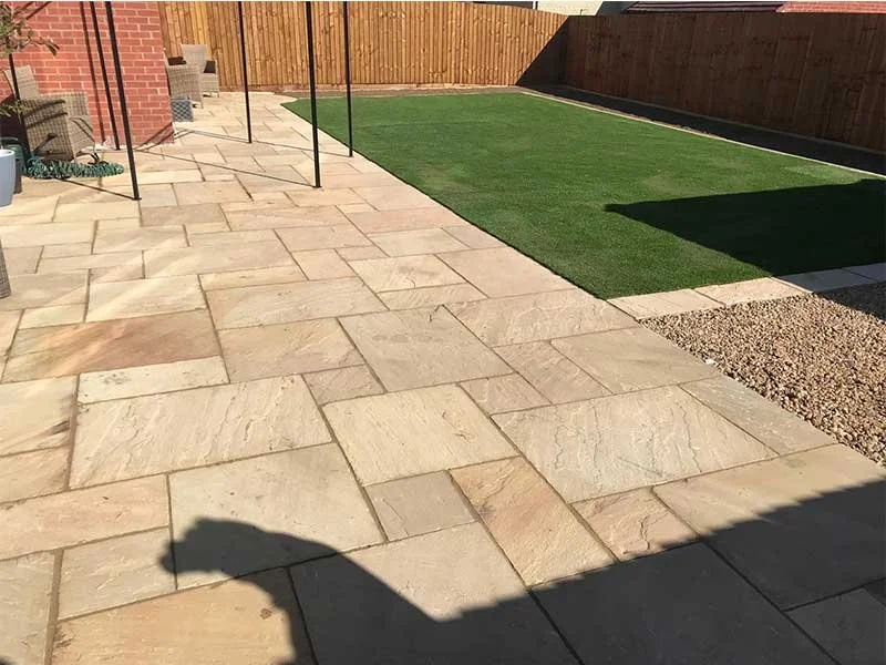 A backyard patio with large beige stone tiles, a patch of artificial grass, a wooden fence, and some gardening tools and plants on the patio.