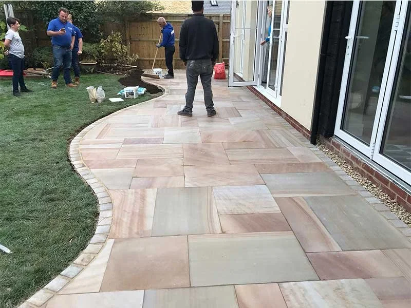 People working on a new stone patio with a curved edge in a backyard, next to a house with sliding glass doors.