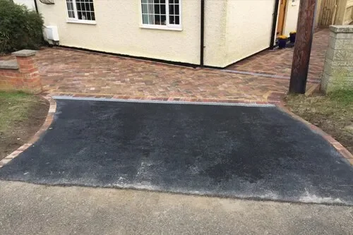 A freshly paved driveway with black asphalt, bordered by brick and stone pavers, leading to a house with a cream-colored exterior.