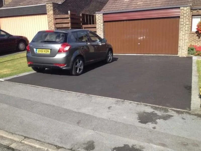 The image shows a freshly paved driveway with a gray hatchback car parked on it. The driveway is bordered by a residential house with a brick wall and a brown garage door.
