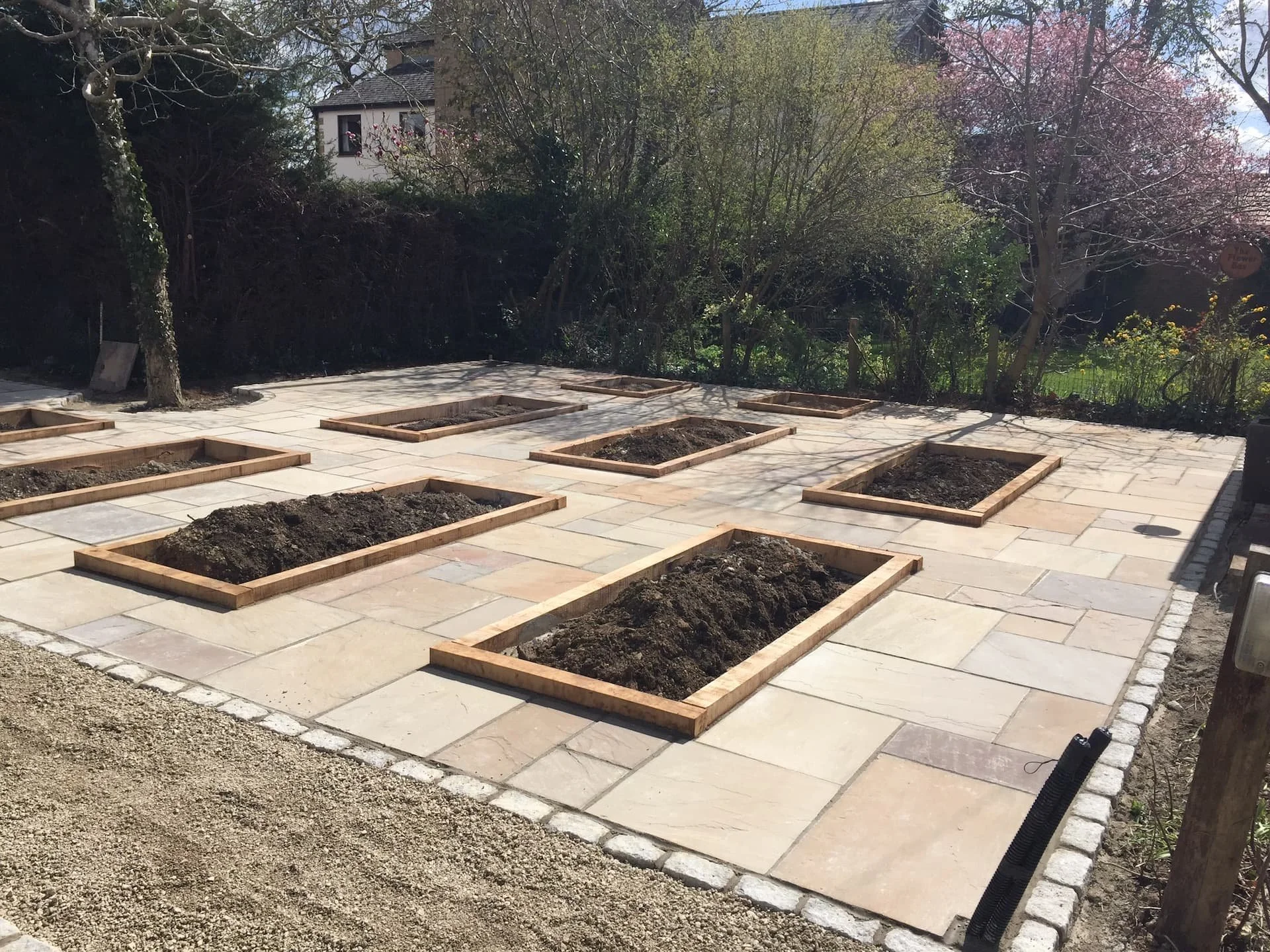 Backyard patio with square garden beds outlined in wood, some filled with soil, surrounded by paving stones, with trees and a house in the background.