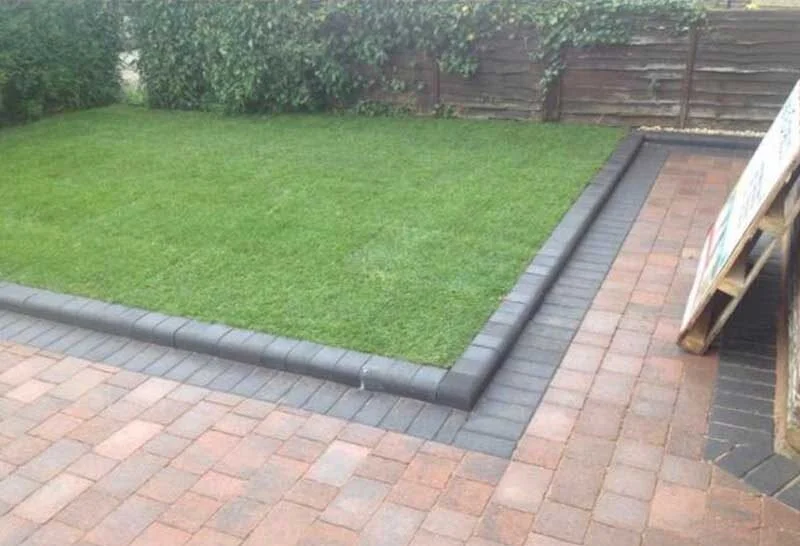 A landscaped backyard showing a small patch of green grass enclosed by black bricks, with a brick patio area surrounding it and a wooden fence in the background.