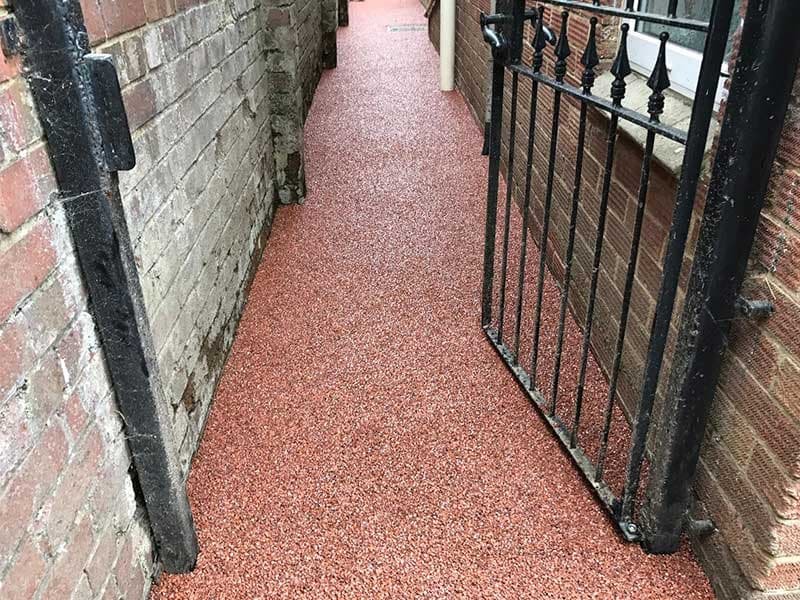 Narrow outdoor alleyway between brick buildings with a red textured floor and a black metal gate on the right side.
