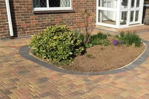 A small garden bed with green bush, purple flowers, and soil, bordered by bricks, next to a brick house with a window and a white porch.