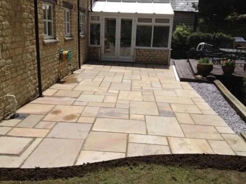 Freshly laid stone paving on backyard patio, adjacent to brick house with glass conservatory, and a raised wooden deck with potted plants.