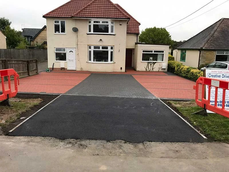Newly paved driveway and patio in front of a two-story house with a red tiled roof, white walls, and multiple windows. Construction barriers are on both sides of the driveway.