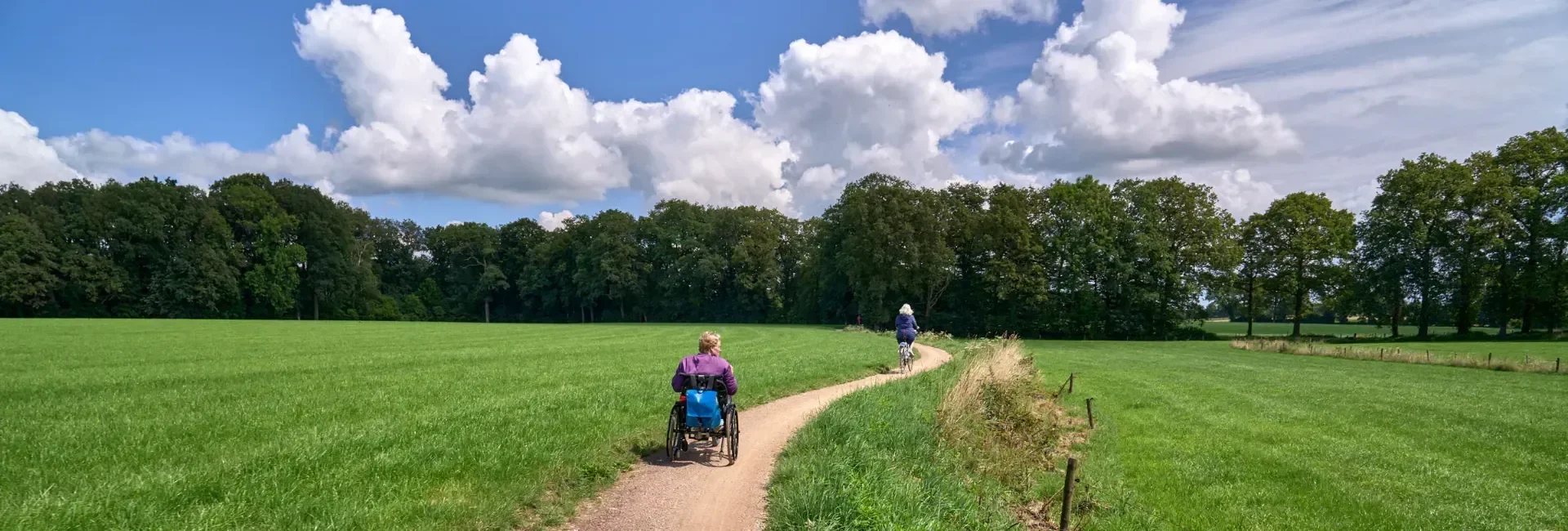 Twee mensen fietsen en een persoon in een rolstoel rijden op een smal pad door een groen veld onder een blauwe hemel met witte wolken, omringd door bomen.