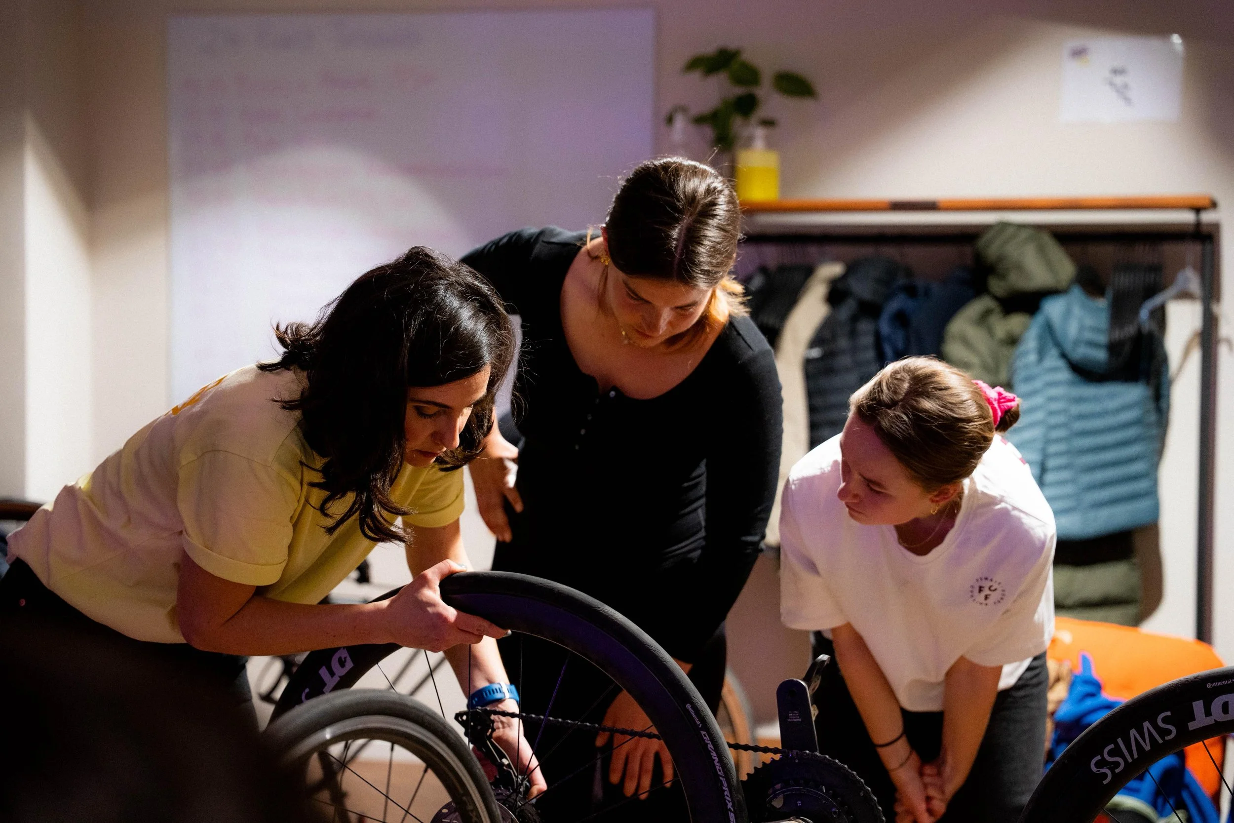Drei Frauen schauen gemeinsam auf ein Fahrrad, das auf einem Rollstuhl montiert ist, in einem Raum mit Jacken an der Garderobe.