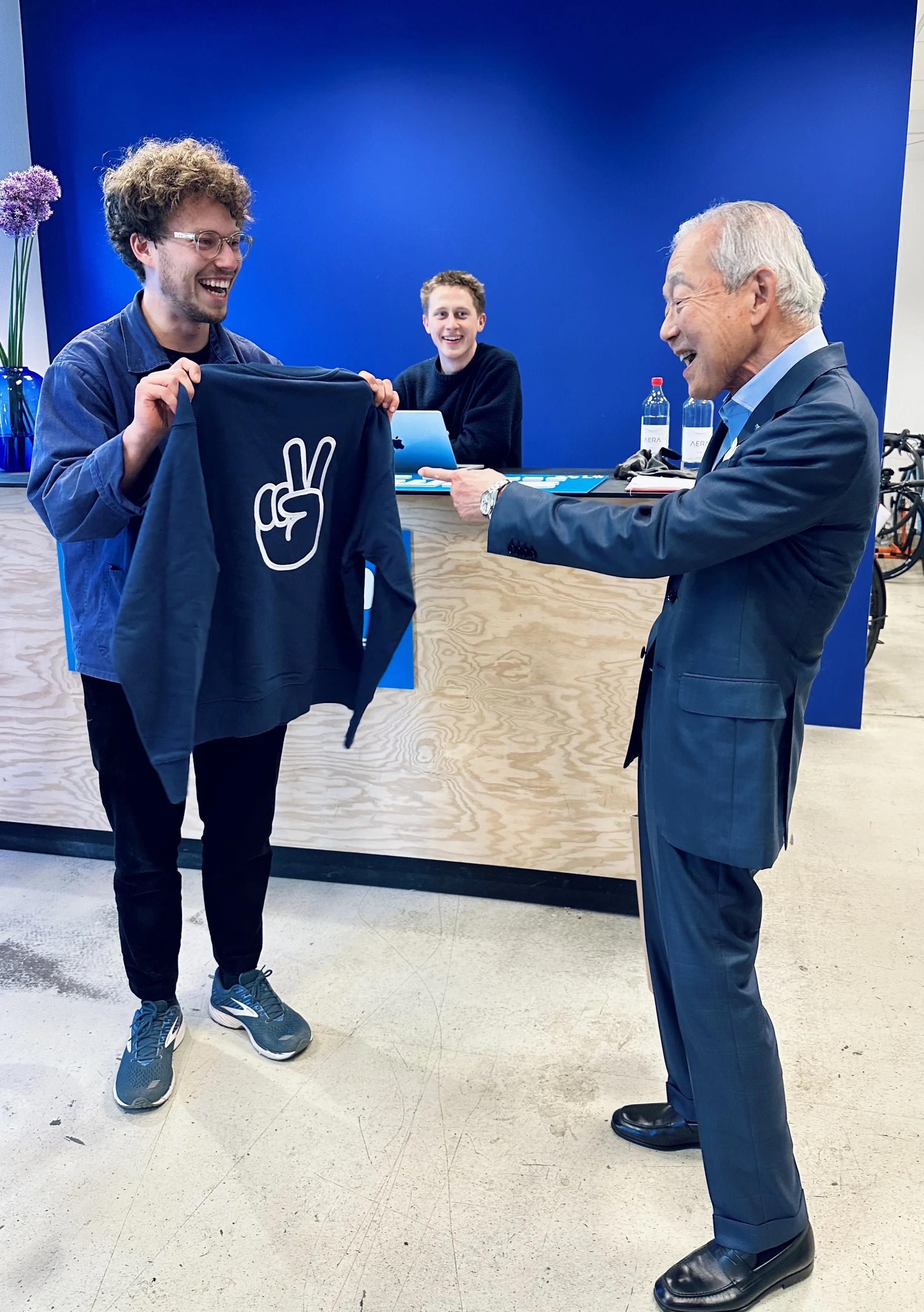 Zwei Männer, einer in einem Anzug, der anderen in einer Freizeitjacke, lachen und tauschen Geschenke aus, während im Hintergrund ein junger Mann an einem Tisch sitzt, alle vor einer blauen Wand.