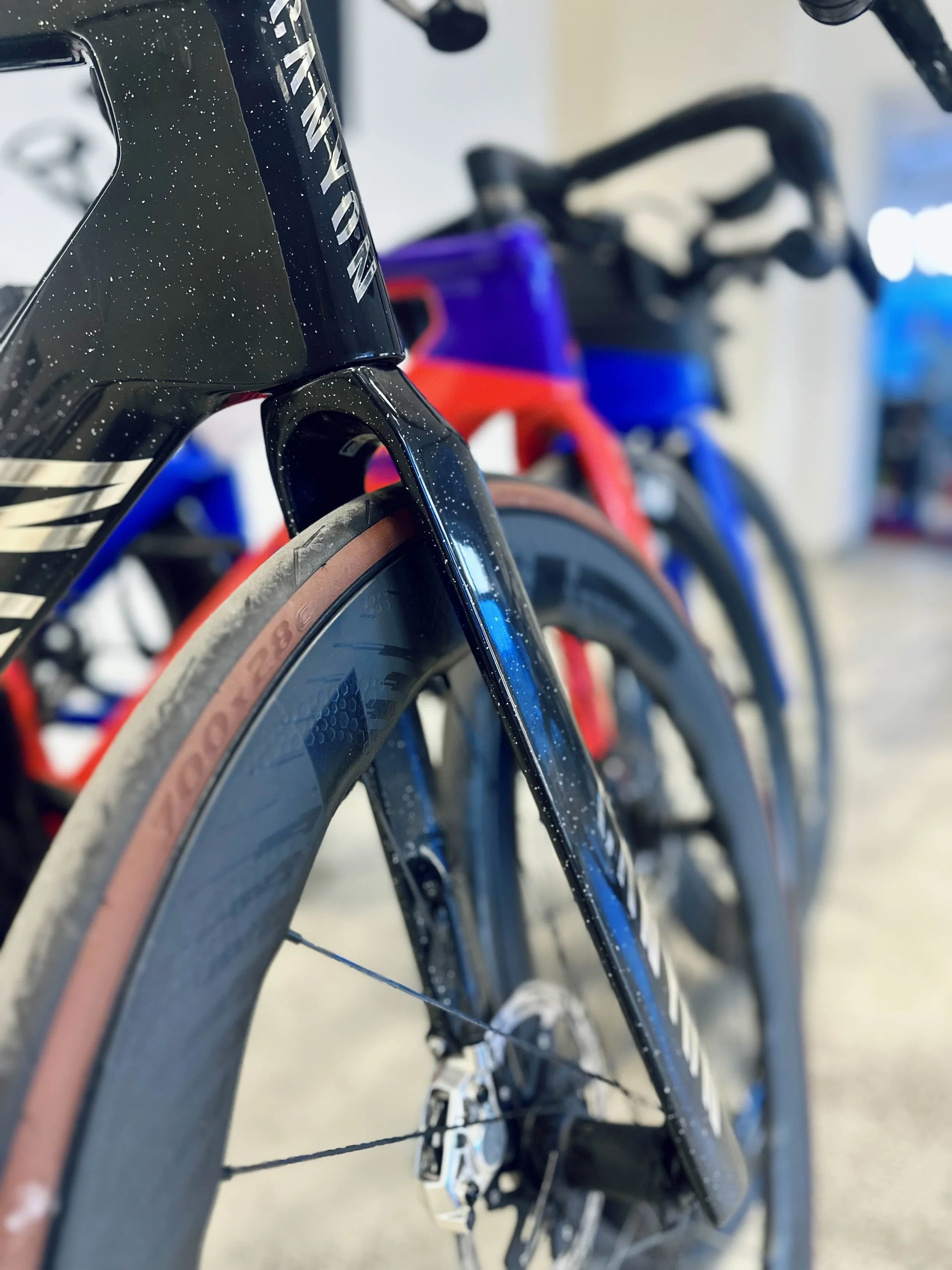 Nahaufnahme von einem modernen Rennrad mit schwarzem Rahmen, braunen Reifen und blauen und roten Akzenten, im Hintergrund weitere Räder sichtbar.