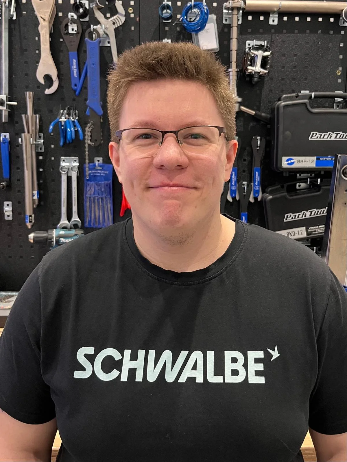 Ein Mann trägt ein schwarzes T-Shirt mit weißen Buchstaben, die das Wort 'SCHWALBE' bilden, vor einer Werkzeugwand.