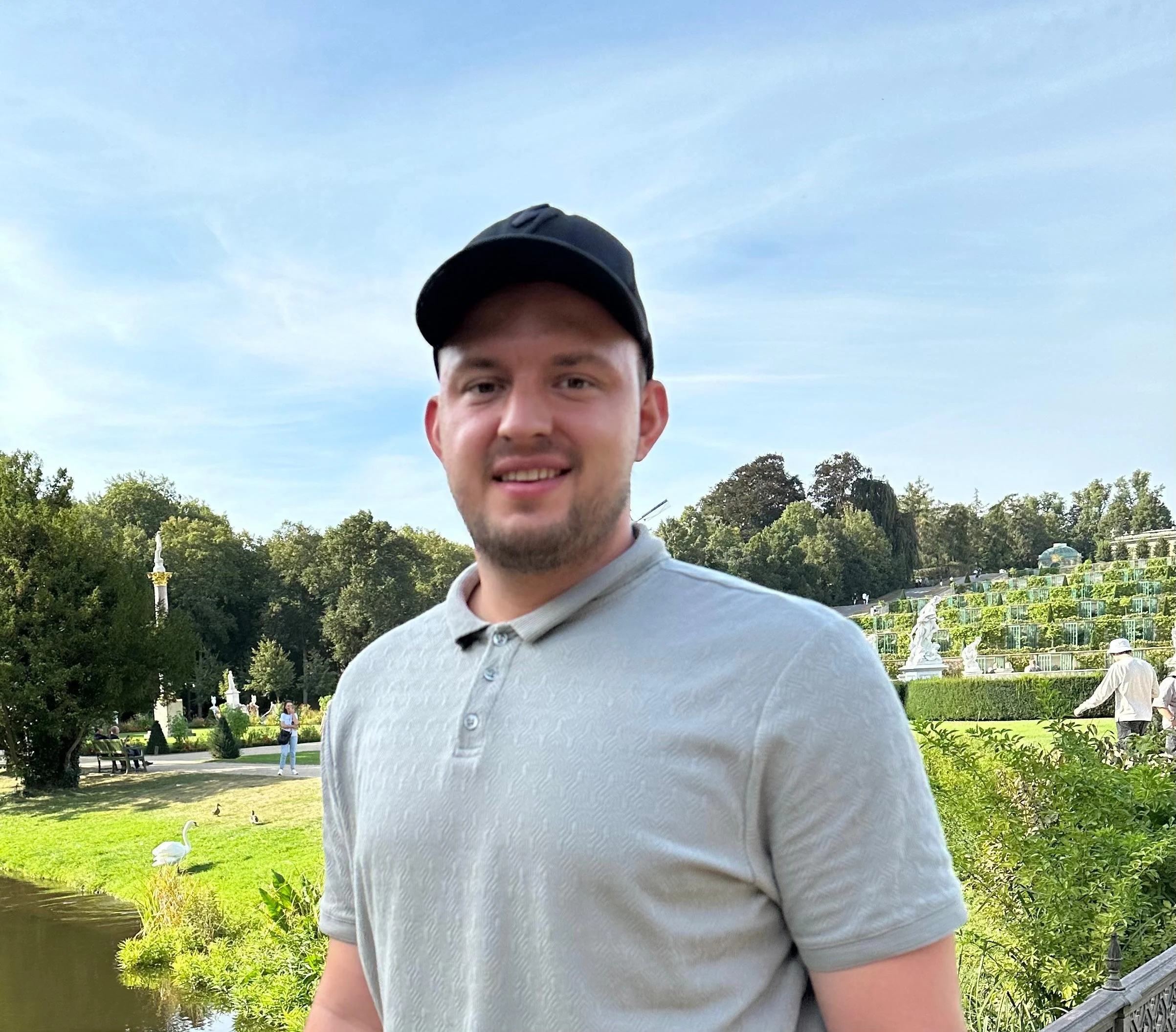 Junger Mann mit grauem Polo-Shirt und schwarzem Cap in einem Park mit Wasser, weißen Schwänen, Bäumen und einer Treppe im Hintergrund.