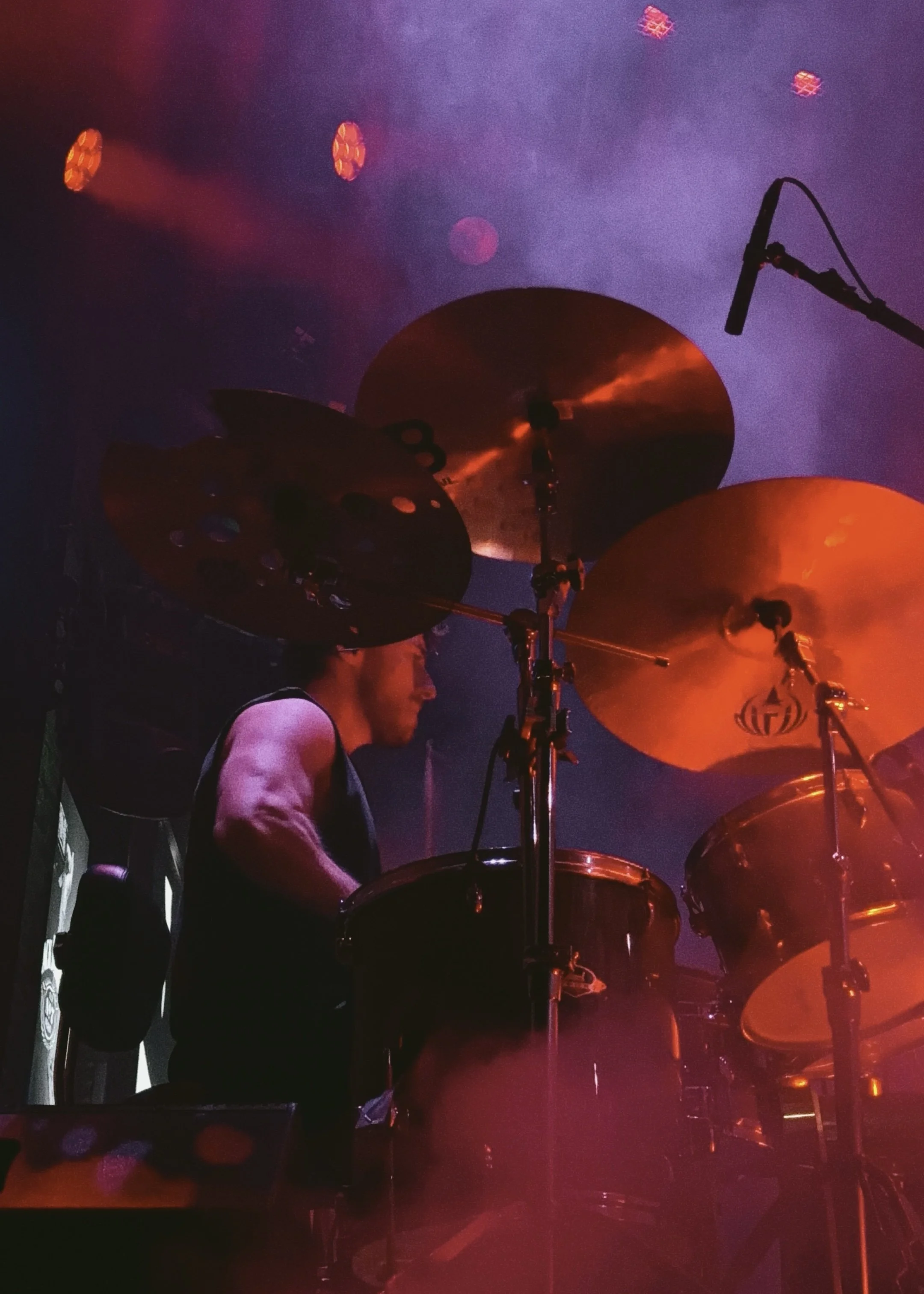 A drummer in a dark setting illuminated by orange and purple stage lights, with visible cymbals, drums, and a microphone.