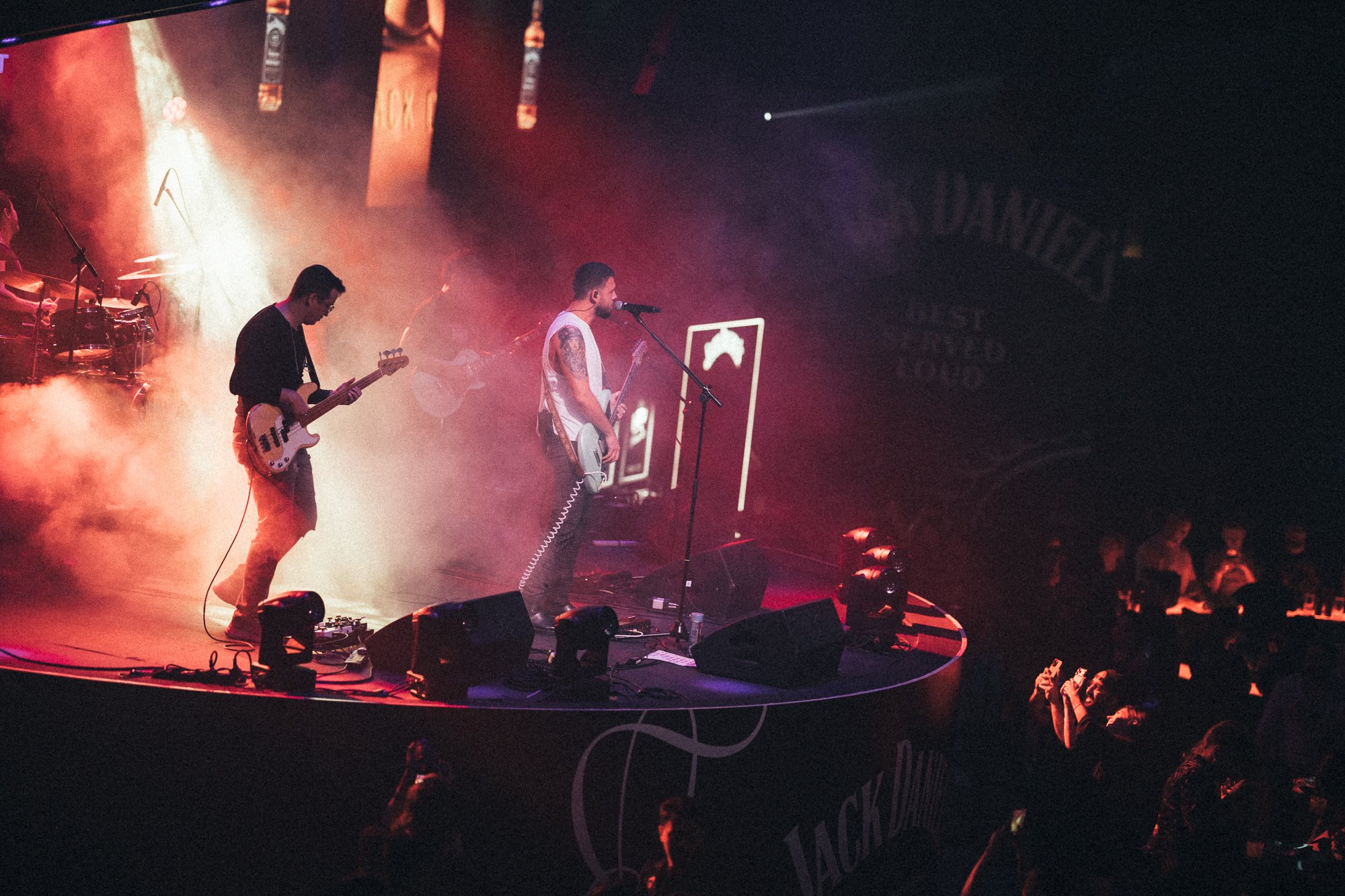A band performs on stage with colorful lighting and smoke effects, with several musicians playing guitars and drums, and audience visible in front.