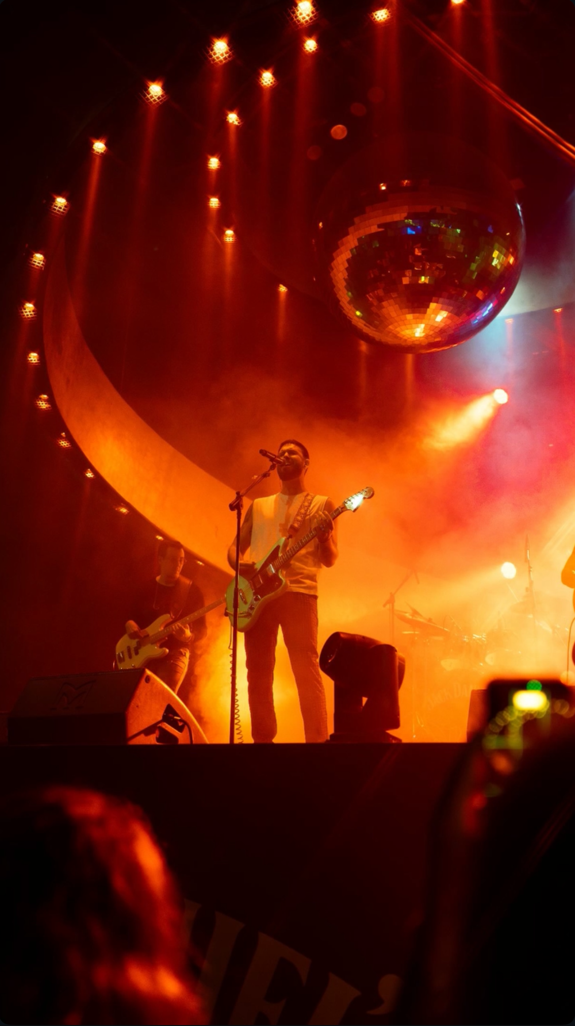 A band performs on stage with orange and yellow lighting. A male singer with an acoustic guitar stands at the center singing into a microphone. Other band members are playing instruments nearby. A large disco ball hangs from the ceiling, reflecting colorful lights.