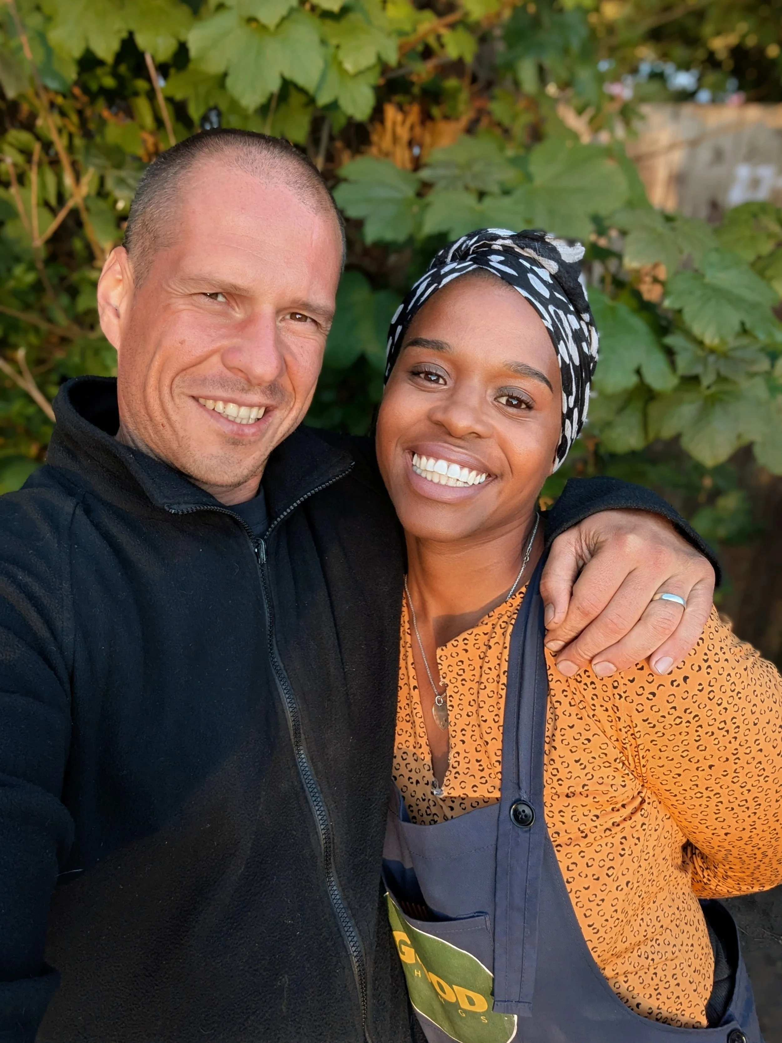 A smiling man and woman taking a selfie outdoors with green foliage in the background. The man is wearing a black zip-up jacket, and the woman is wearing an orange patterned shirt, a black and white headscarf, and an apron.