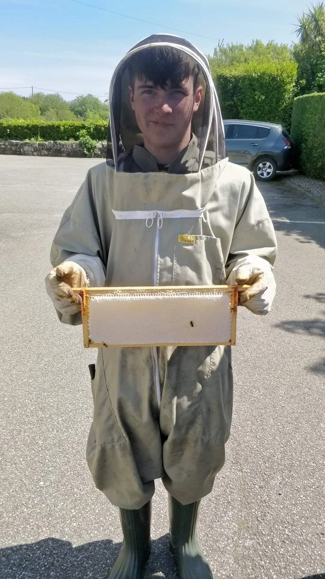 Person dressed in a beekeeping suit holding a honeycomb frame outdoors.