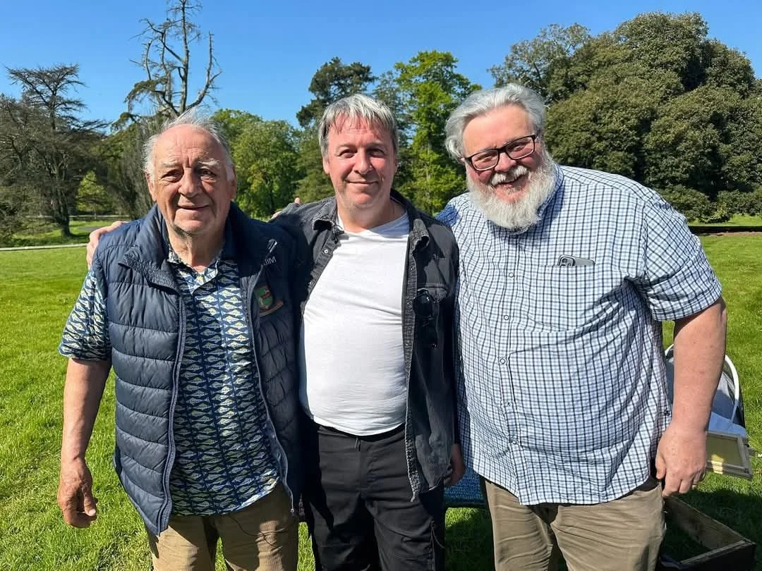 Three middle-aged men standing together outdoors in a park, smiling at the camera. The background shows green grass, trees, and a clear blue sky.