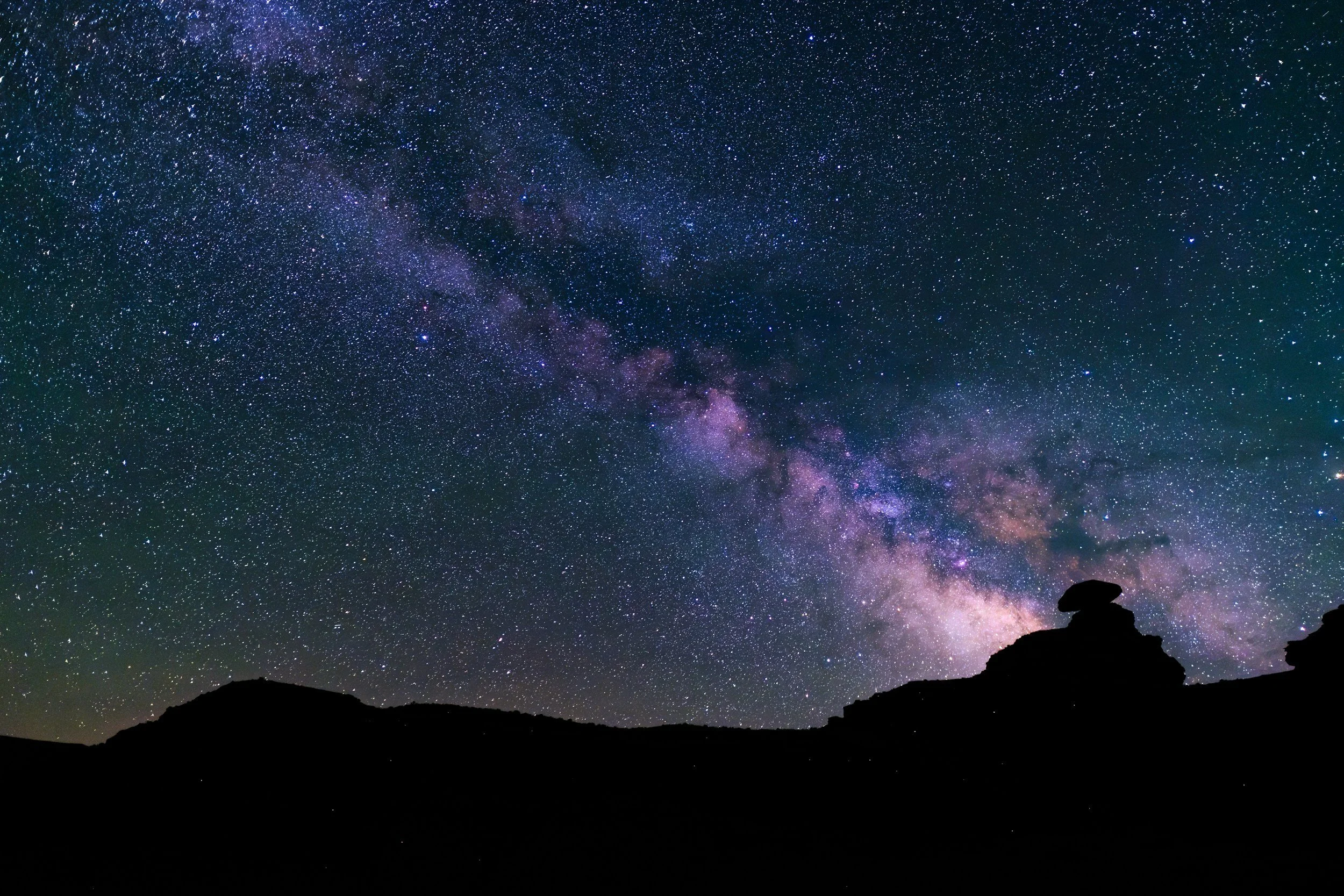 Night sky filled with stars and the Milky Way galaxy, with a silhouette of a rocky hill or mountain in the foreground.
