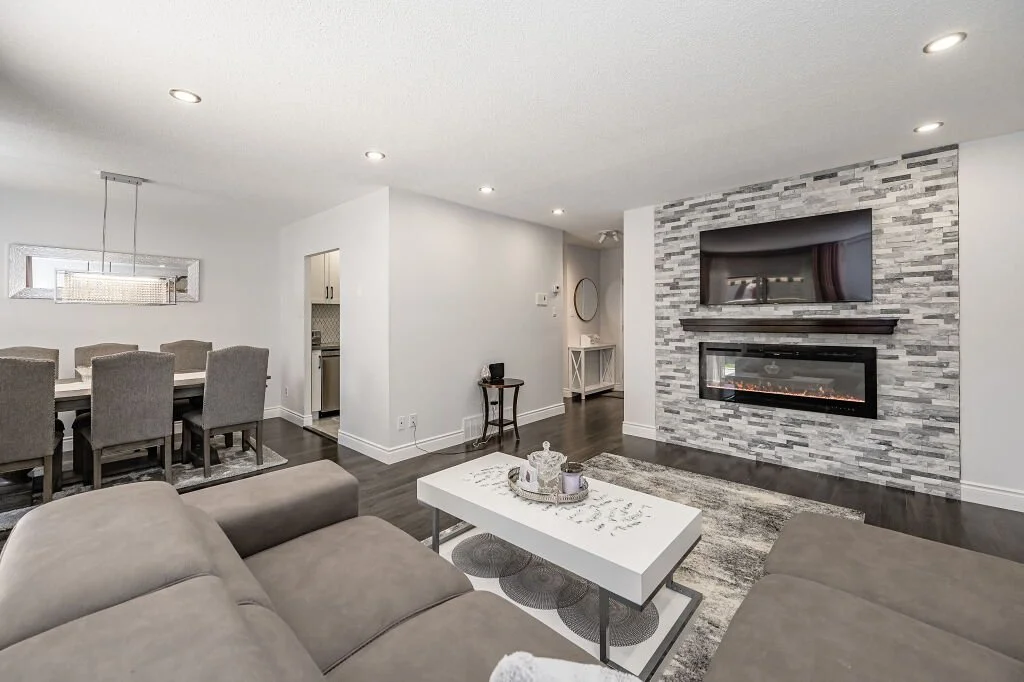 Living room with a beige sectional sofa, white coffee table, on a gray rug, and an accent wall with a mounted TV and electric fireplace. Dining area with a rectangular table and eight gray chairs, and a view into the kitchen.