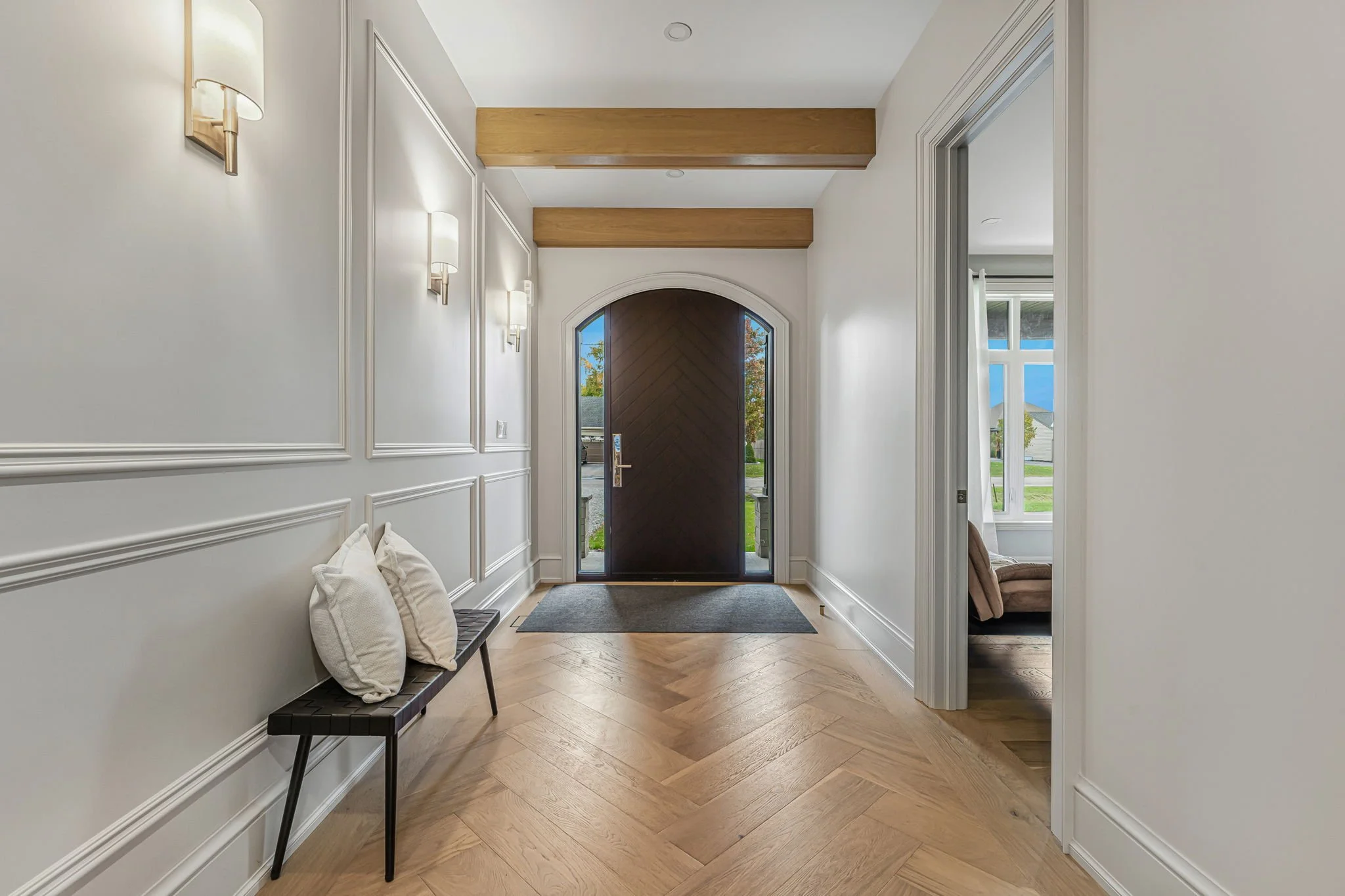 Entryway with wooden flooring, white walls with decorative molding, wall-mounted lights, a dark wooden front door with glass panes on sides, a small rug in front of the door, and a bench with two white pillows on it. An opening shows a room with a window and a brown armchair.