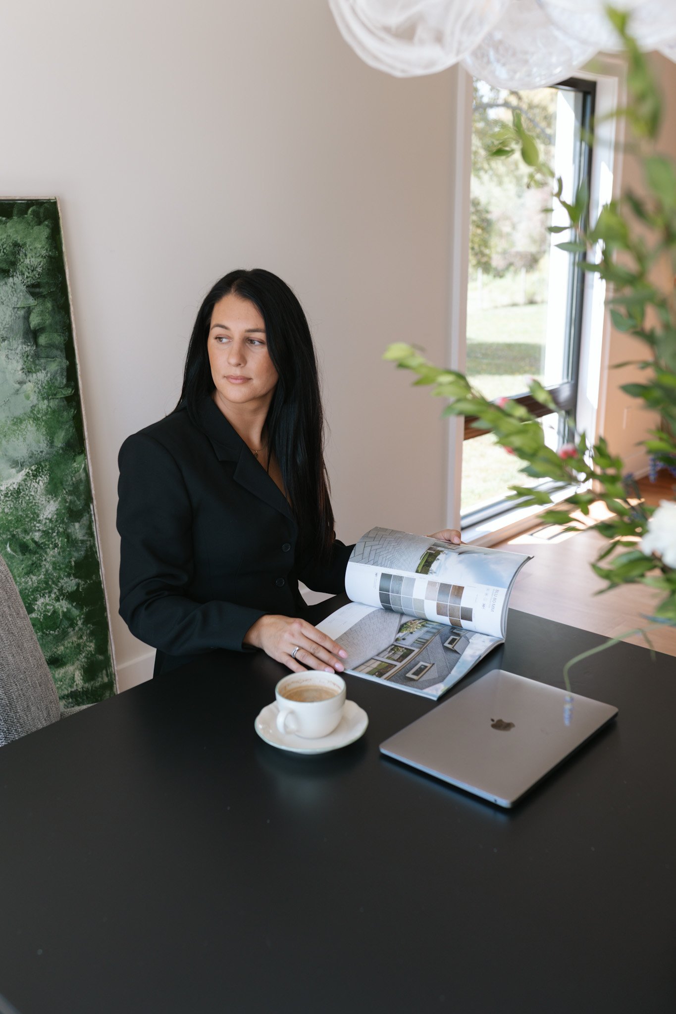 A woman with long black hair, wearing a black blazer, sitting at a black table, looking to her left while reading a magazine. There is a cup of coffee on a saucer and a closed silver laptop on the table. The setting includes a window with natural light and some green plants.