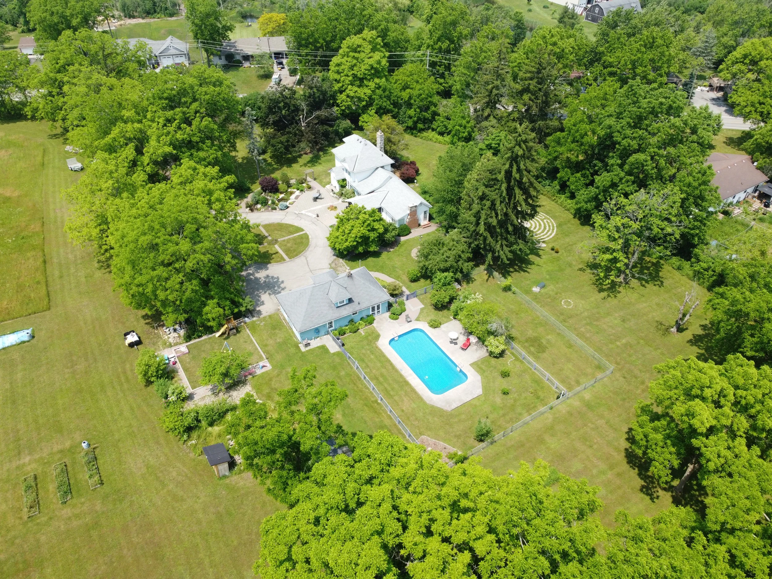 Aerial view of a large backyard with a swimming pool, surrounded by trees, with a house and a smaller outbuilding.