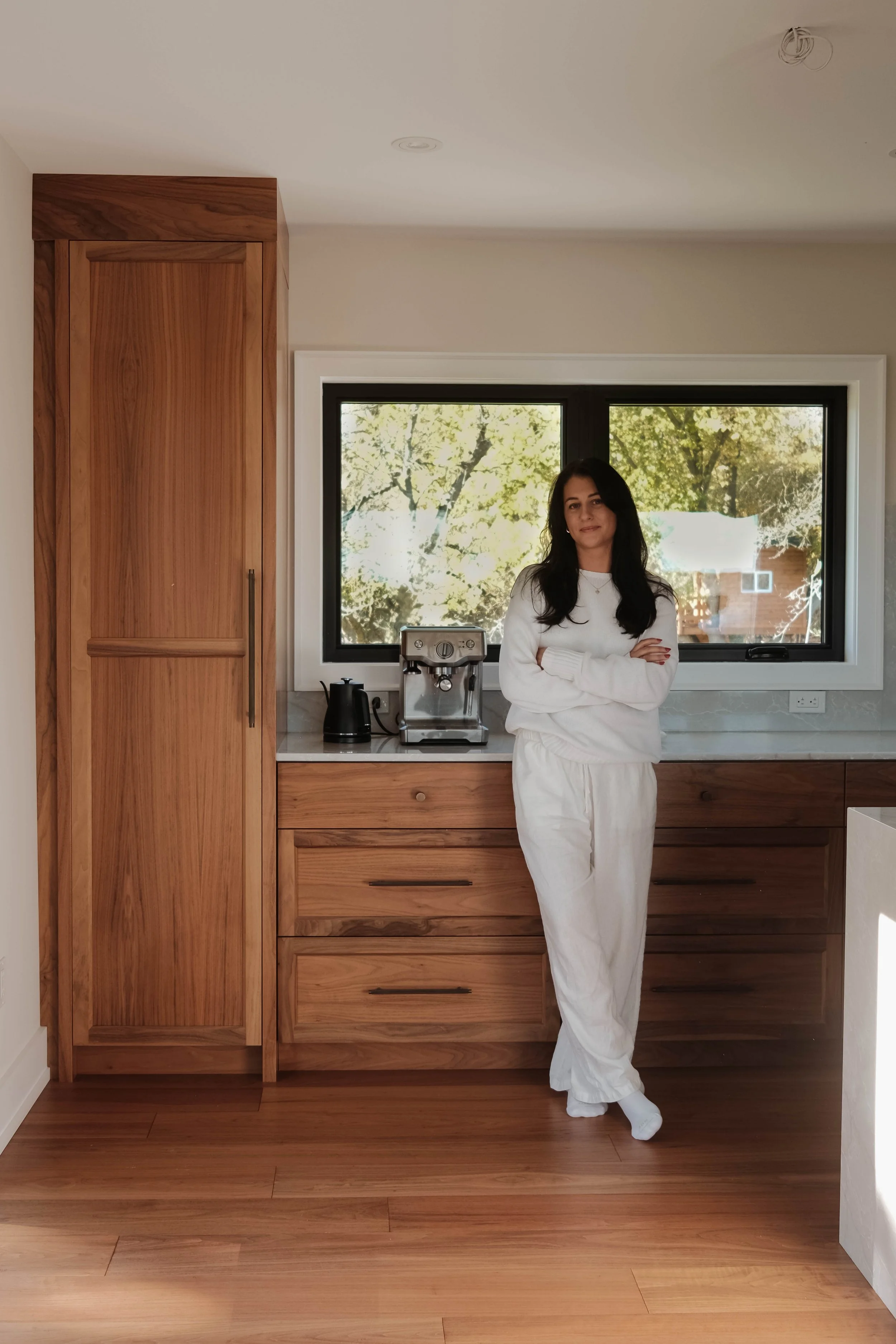 Woman with dark hair wearing white pajamas standing in a modern kitchen with wooden cabinets, a black kettle, and a coffee machine, in front of a large window showing trees outside.
