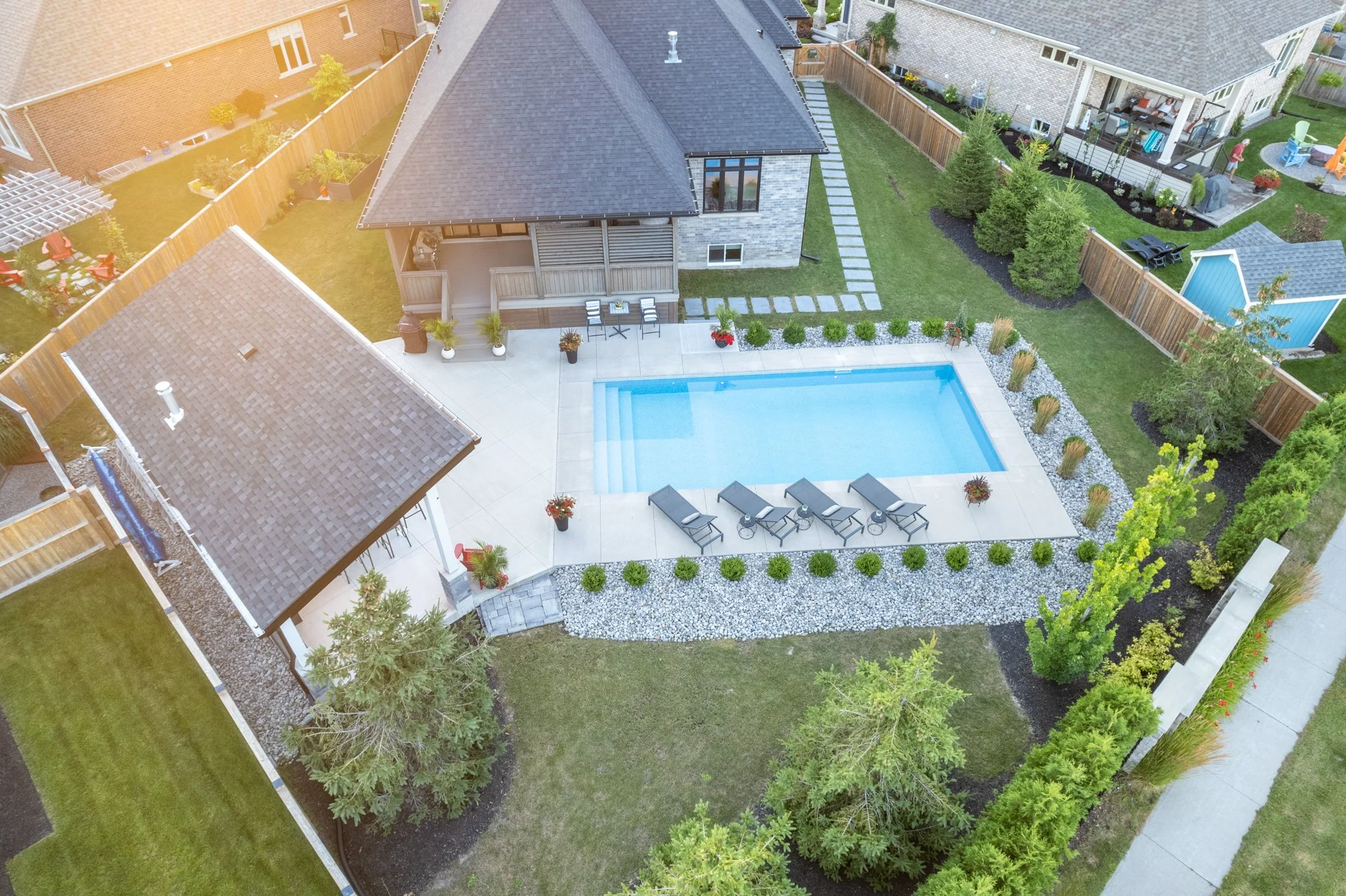 Aerial view of a backyard with a swimming pool, surrounded by a patio, garden beds, and trees; a house with a porch and gabled roof nearby.