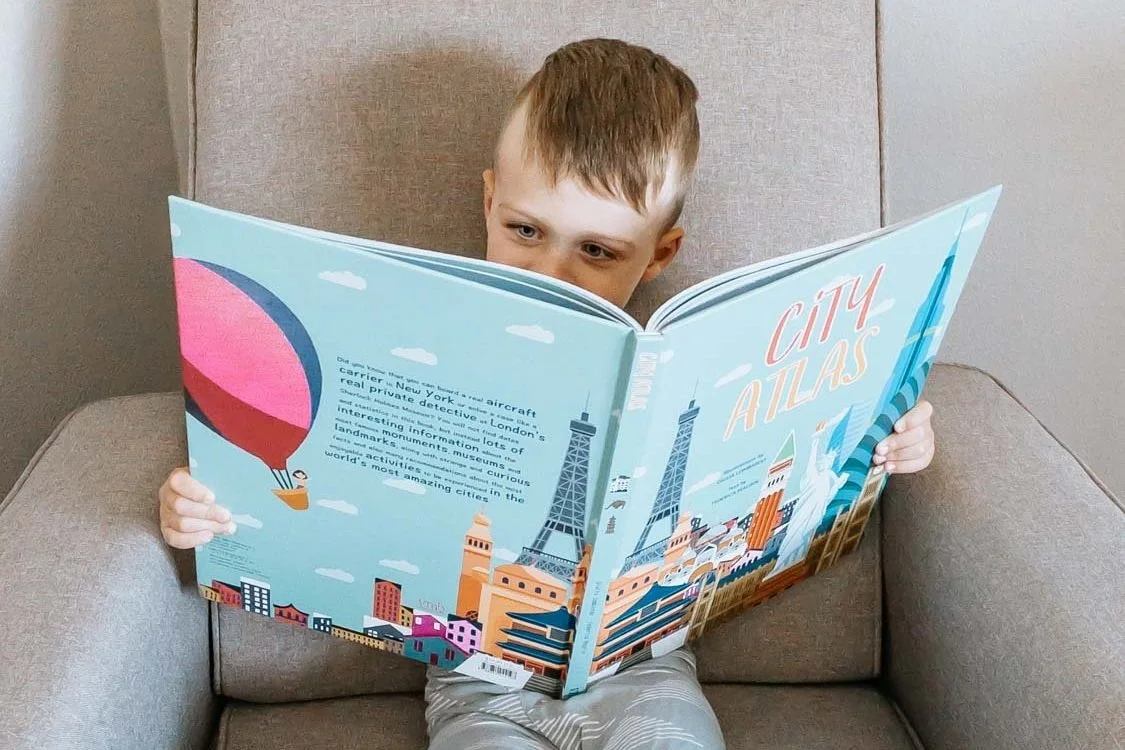 Little boy sitting in chair reading a kids city atlas of places to visit on future family trips