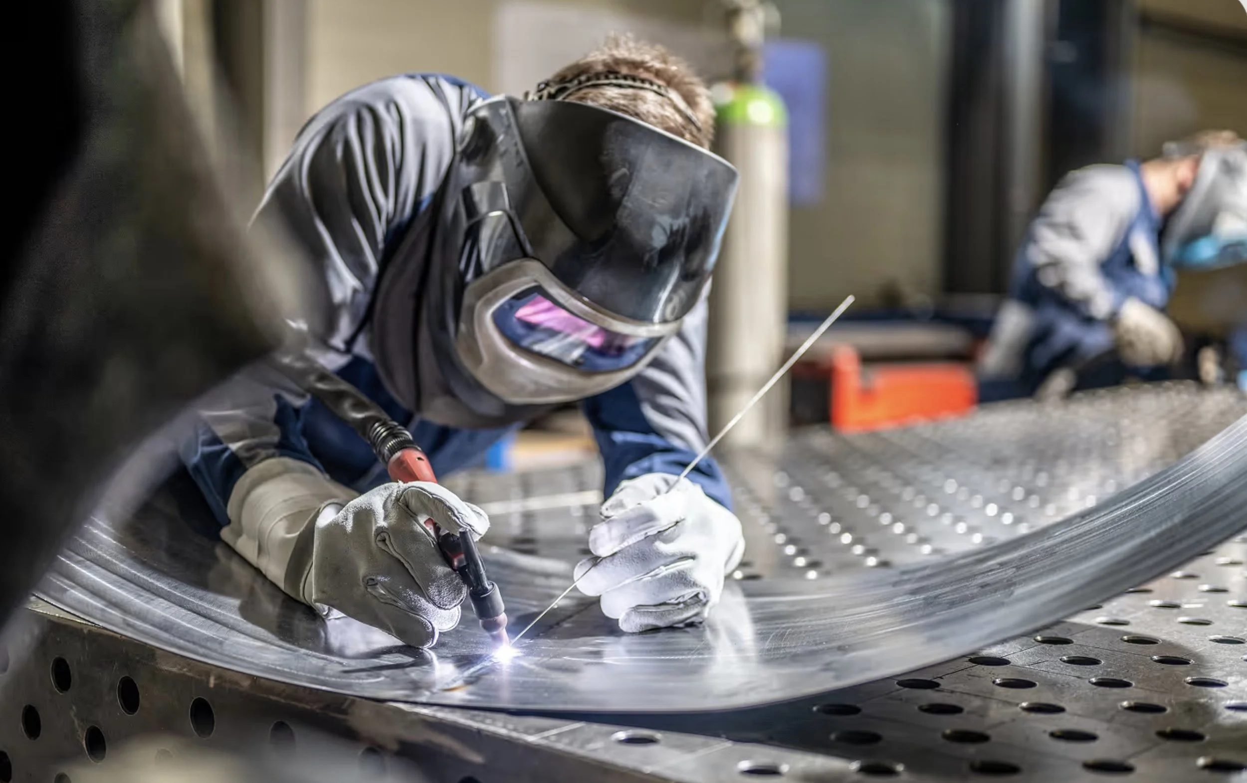A machinist is welding two pieces of metal with his protective face shield and gloves.