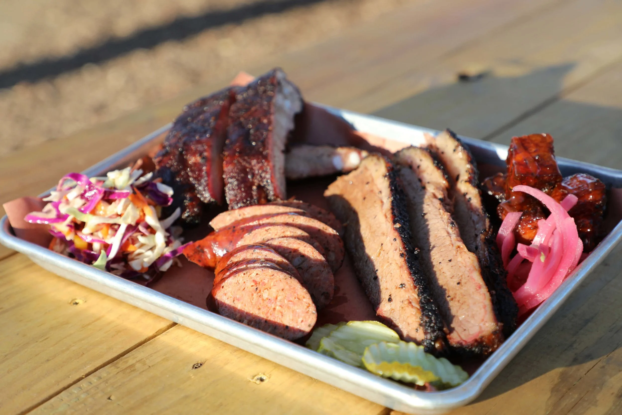 Barbecue platter with sliced brisket, sausage, ribs, coleslaw, pickles, and pickled onions