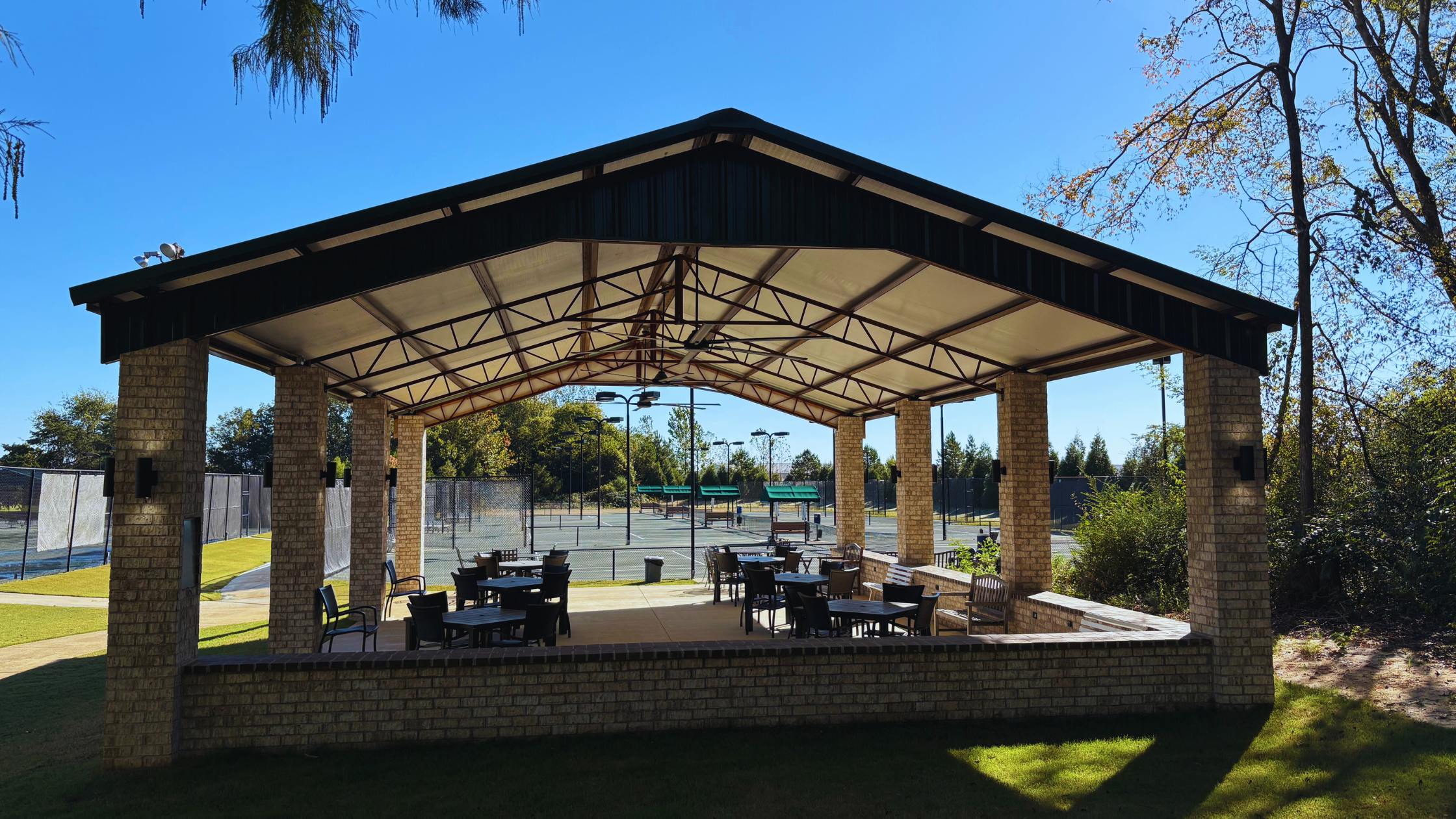 Pole barn pavilion structure supplied by Phoenix Building Solutions in Tupelo, MS, featuring a metal roof and exposed steel trusses overlooking tennis courts.