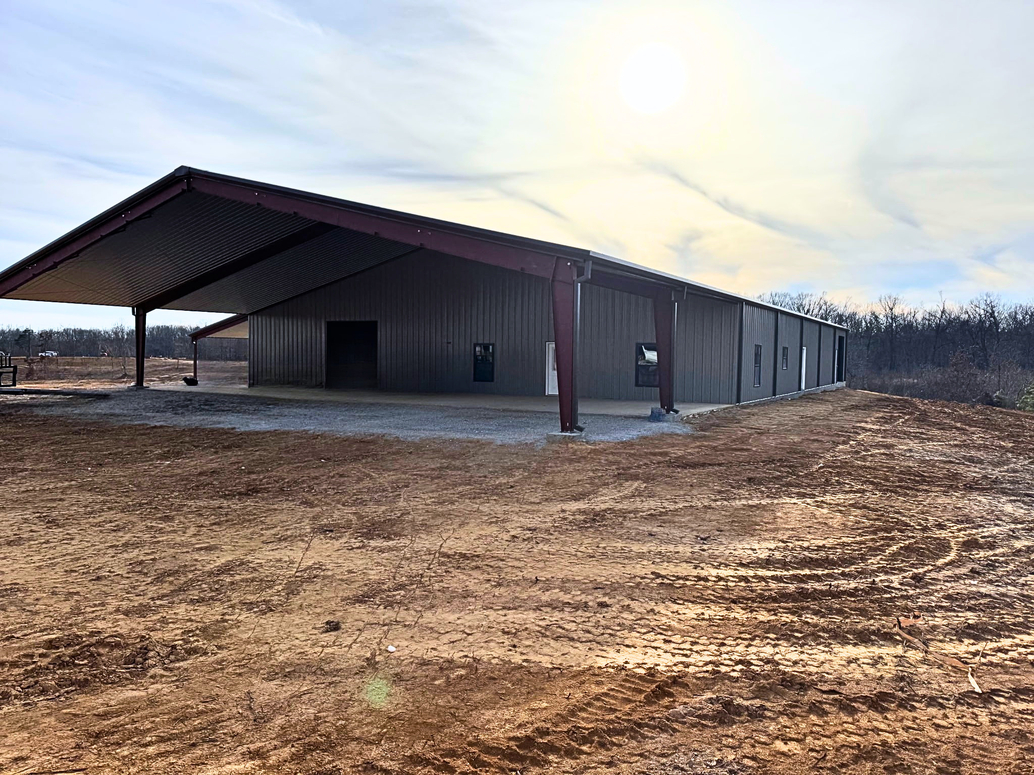 A wide-angle perspective of a large, modern pre-engineered metal building under a bright sky. The structure features dark grey corrugated metal siding and a deep red steel frame. A massive integrated overhang creates a shaded outdoor workspace or sto