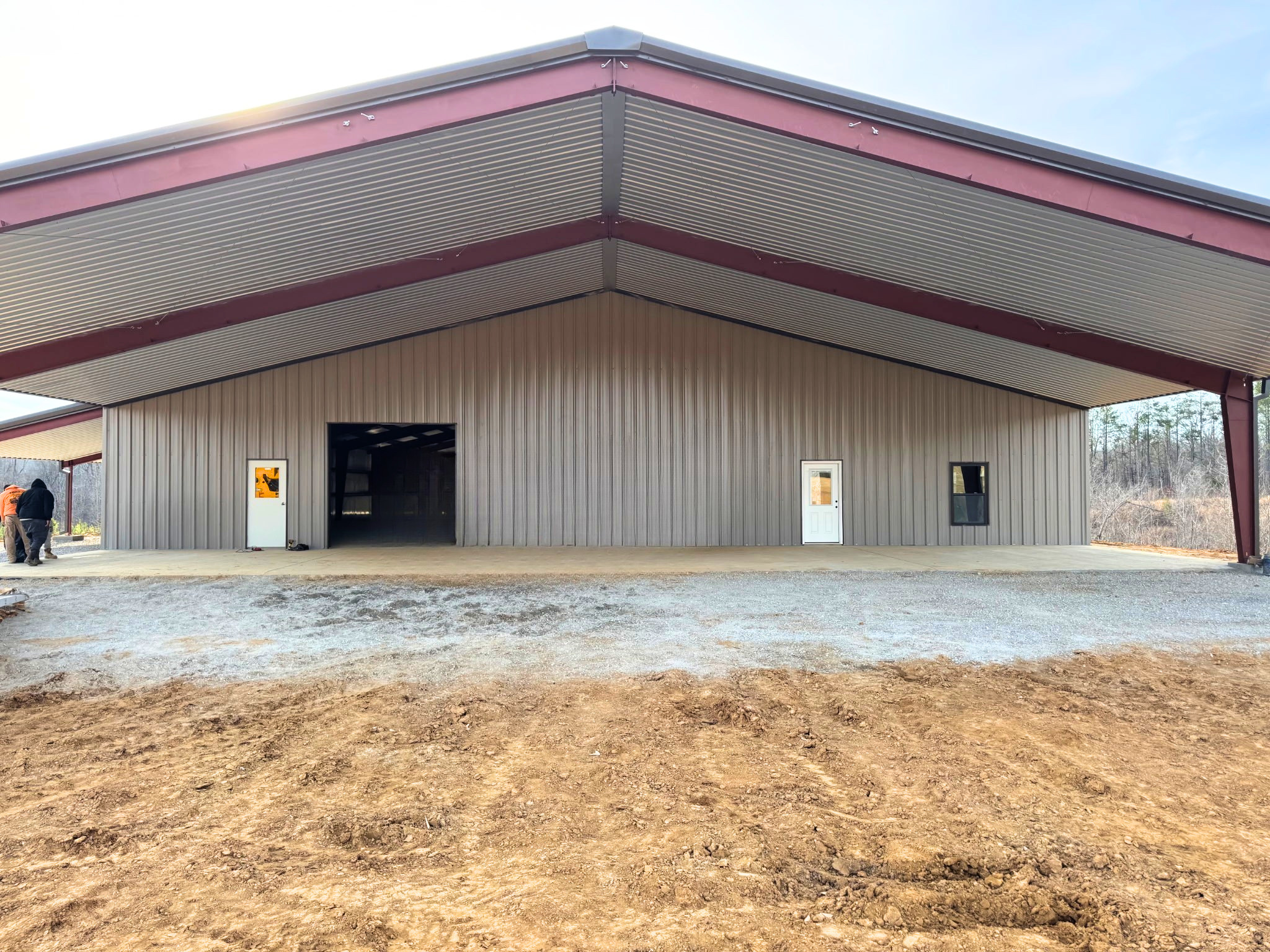 A symmetrical, straight-on view of the building's gable end. The wide red steel rafters support a large covered porch area. The main wall features a central open bay door flanked by two white pedestrian doors and a single window. The foreground shows