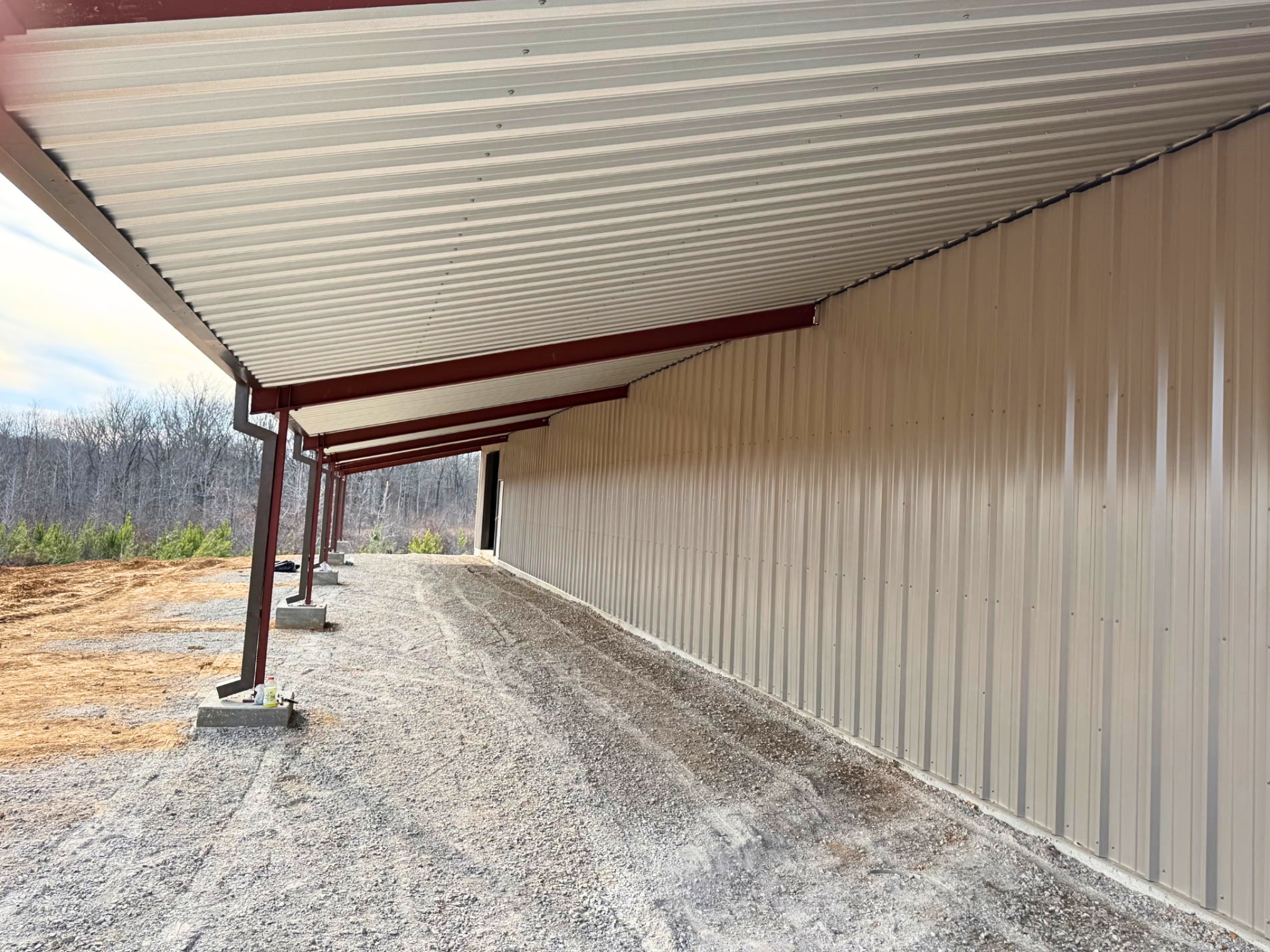 A view from beneath the building’s side overhang, looking down the length of the structure. The red steel I-beam rafters and the underside of the ribbed metal roofing are visible. The exterior wall is tan corrugated metal, and the support columns are