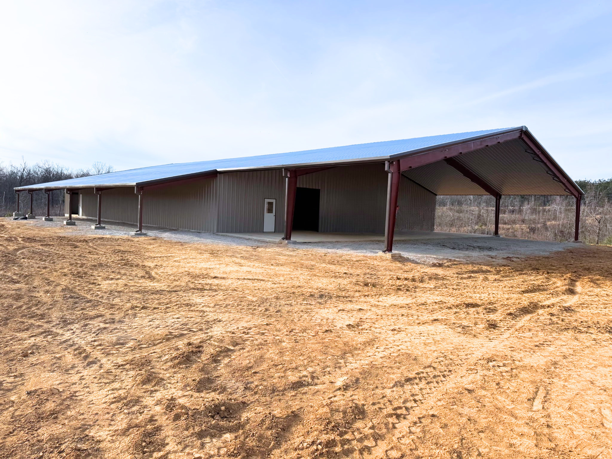 A side-view of the long metal building showing a continuous lean-to roof extension supported by red steel columns. The building has a low-sloped roof and tan-grey vertical metal wall panels. The structure sits on a concrete foundation in a rural, cle