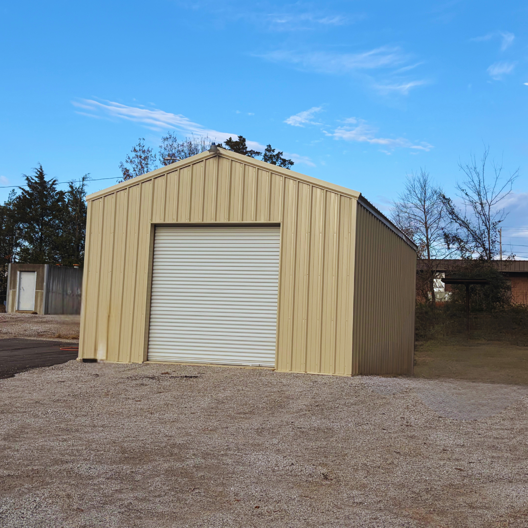 A small, standalone tan metal utility enclosed pole barn with a single white roll-up garage door centered on the front gable by Phoenix Building Solutions. - Mississippi | Metal Buildings | Barndominiums | Metal Roofing | Pole Barns
